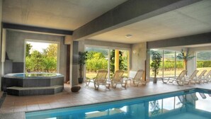 Una piscina interna con poltrone a sdraio e vasca idromassaggio, circondata da ampie finestre con vista su un'area verde.