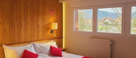 A hotel room with a wooden headboard, a bed with white bedding and red accents, a window with a view of greenery, and a small stool.