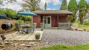 Un petit bâtiment en bois avec une porte, une terrasse avec une table et des chaises, un barbecue et un chemin de gravier.