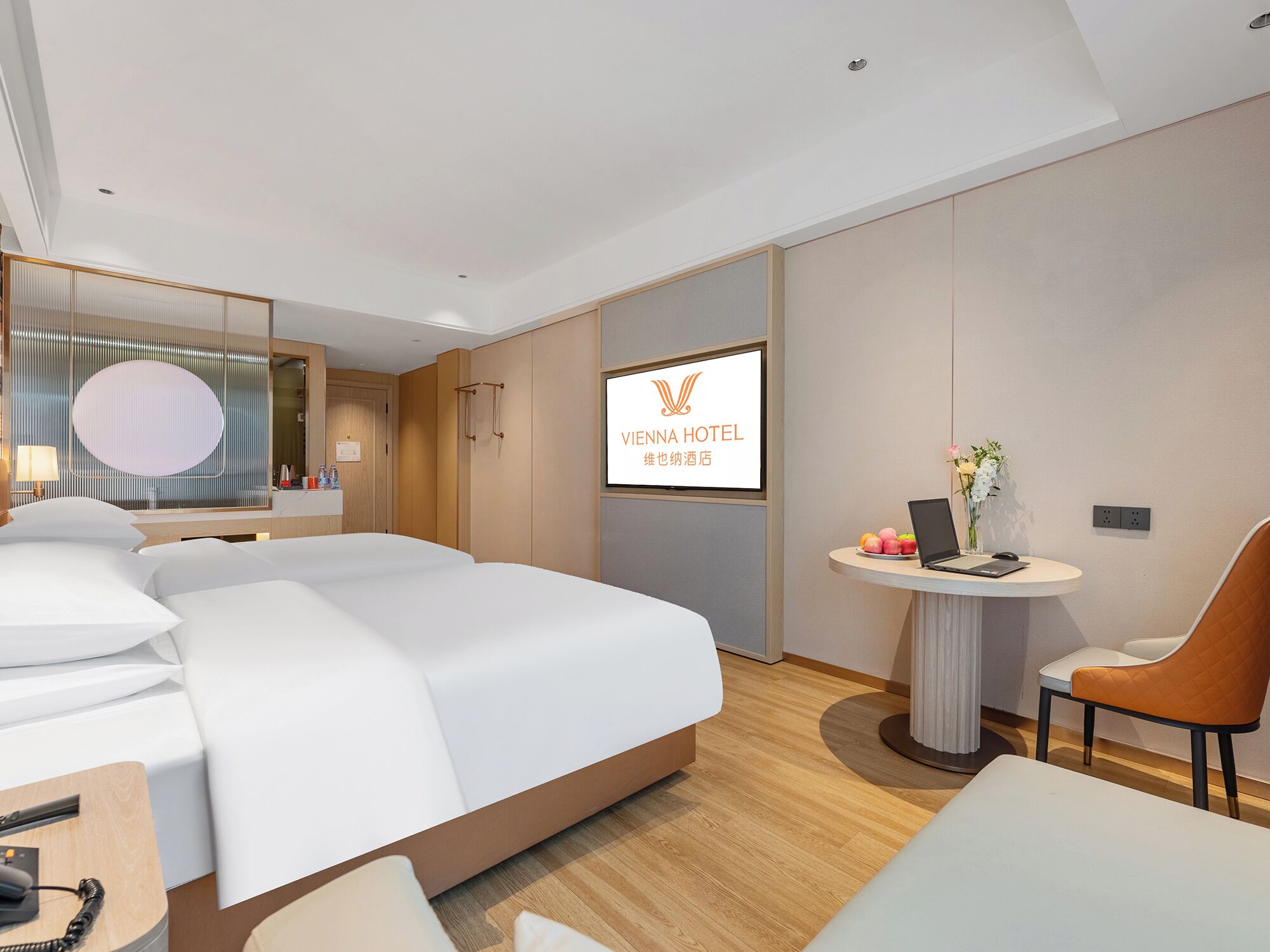 Una camera d'hotel con un letto, una scrivania con il telefono, una sedia, un tavolino con un portatile e fiori, e un televisore che mostra il nome dell'hotel.