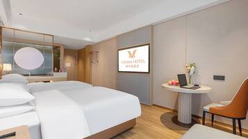 Una camera d'hotel con un letto, una scrivania con il telefono, una sedia, un tavolino con un portatile e fiori, e un televisore che mostra il nome dell'hotel.