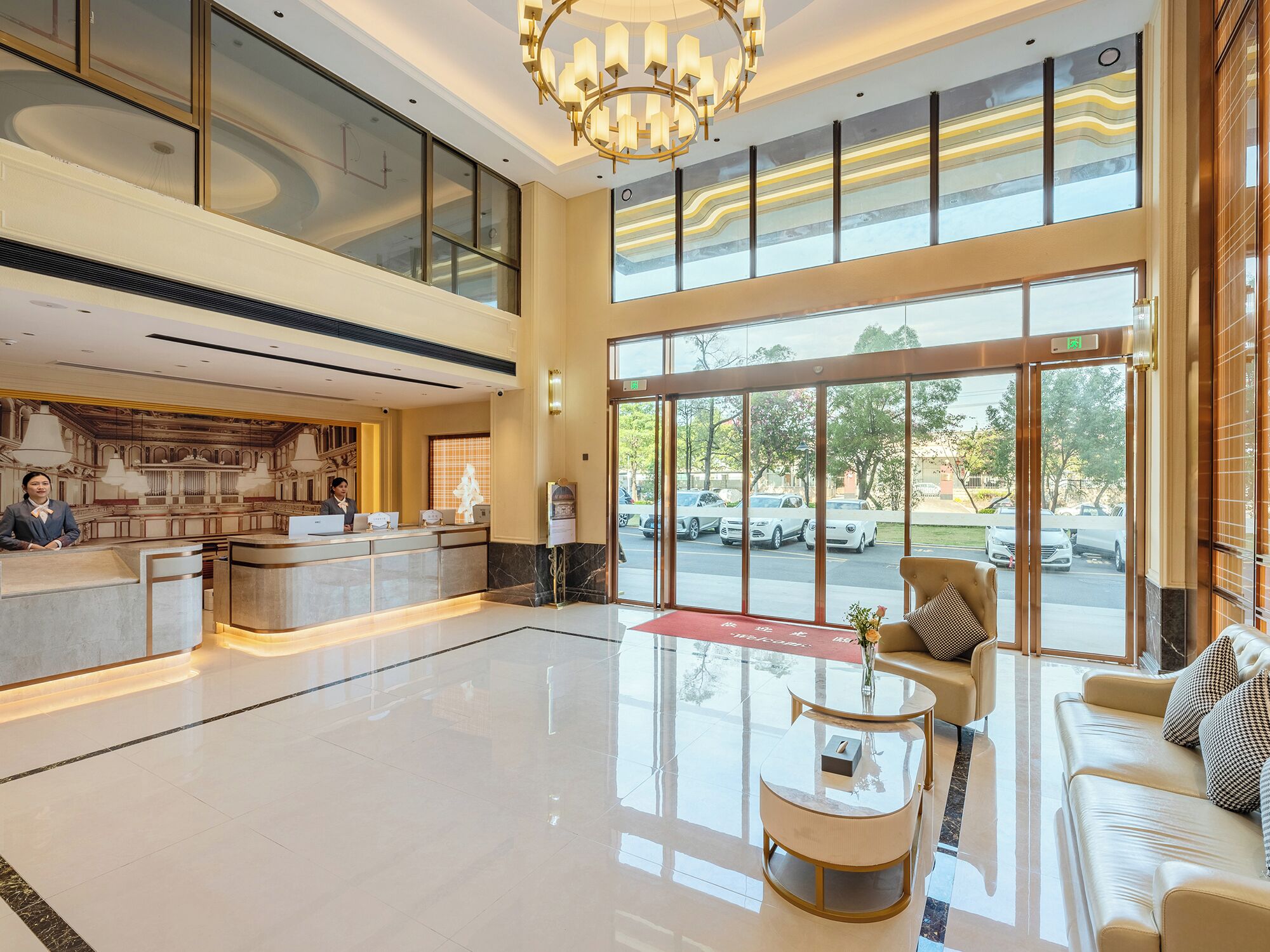 A hotel lobby with a reception desk, seating area, and large windows offering a view of parked cars and trees.