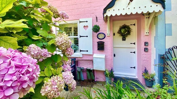 Una cabaña rosa con puerta blanca, una ventana con forma de corazón y una corona en la puerta.