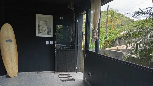 Una habitación con piso de baldosas, una tabla de surf apoyada en la pared, un cuadro enmarcado y una ventana con vista a la vegetación.