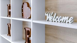 Ein weißes Regal mit Dekorationsobjekten, darunter ein herzförmiges Holzobjekt und ein Schild mit der Aufschrift 'Welcome'.