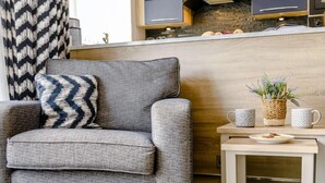 Ein modernes Wohnzimmer mit einem grauen Sessel, einem Holztisch und einer Küchenzeile im Hintergrund.