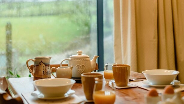 Une table de petit-déjeuner avec des tasses à thé et à café, une théière et un pichet, placée près d’une fenêtre donnant sur un paysage verdoyant.