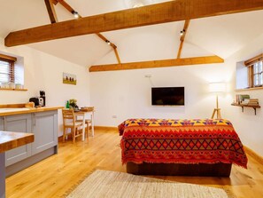 Ein gemütliches Schlafzimmer im Loft-Stil mit Holzbalkendecke, einem Bett mit farbenfroher Decke, einem Schreibtisch mit Stuhl und einem Fernseher an der Wand.