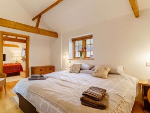Une chambre à coucher avec un lit, des poutres en bois, une fenêtre décorée de fleurs, une table de chevet et une commode.