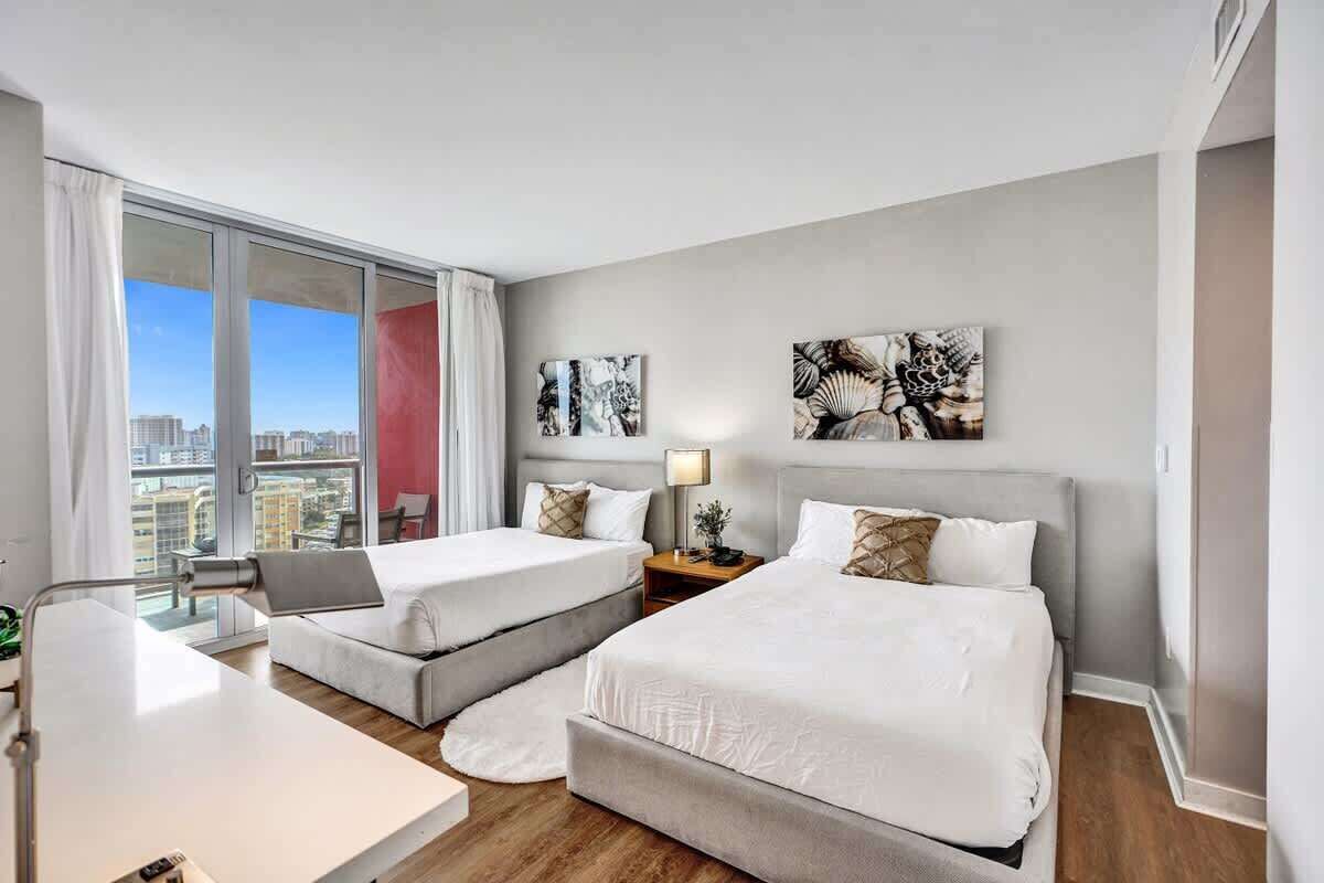 A modern hotel room with two beds, a desk, and a large window offering a city view.