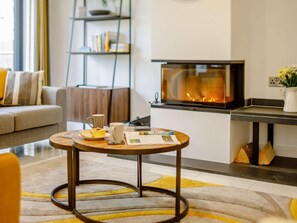 A cozy living room with a fireplace, a sofa, a coffee table with books and a mug, and a bookshelf. - 4 Bed in Bude (oc-89355) (Poughill)