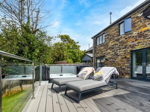 A wooden deck with two lounge chairs, a hot tub, and a stone building with large windows. - 4 Bed in Bude (oc-89355) (Poughill)