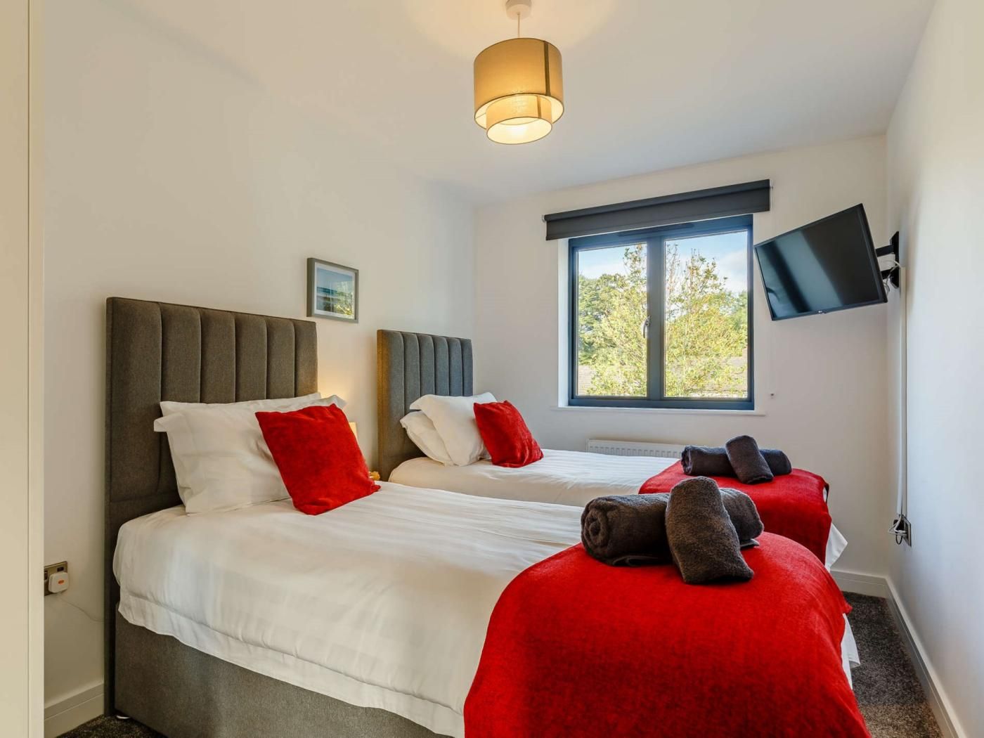 Ein Hotelzimmer mit zwei Betten, einem an der Wand befestigten Fernseher und einem Fenster mit Blick auf Bäume.