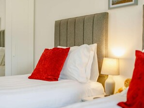A neatly made bed with white and red pillows, a bedside lamp, and a teddy bear on the nightstand. - 4 Bed in Bude (oc-89355) (Poughill)