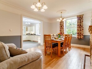 A dining area with a wooden table and chairs, a kitchen in the background, and a view of greenery outside. - 5 Bed in Wisbech (oc-90134) (Wisbech)