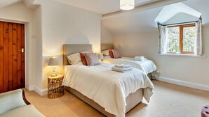 A bedroom with two beds, a wooden door, a round table with a lamp, and a window with curtains. - 3 Bed in Holywell (oc-89614) (Gorsedd)