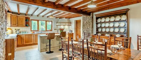A rustic kitchen with wooden beams, a stone wall, and a dining area with a wooden table and chairs.