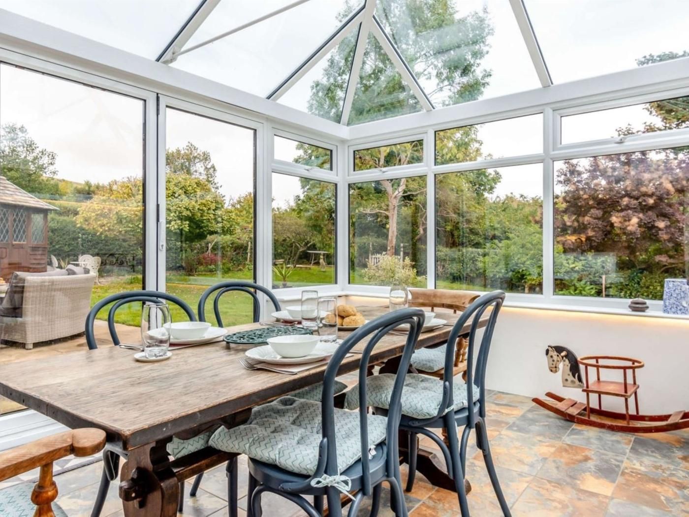 A conservatory with a wooden table and chairs, surrounded by large windows offering a view of a garden.