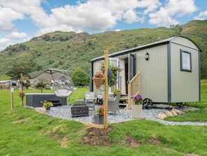 Ein kleines, eingeschossiges Mobilheim mit Veranda, Sitzgelegenheit im Freien und einem Gartenbereich mit Blumentöpfen.