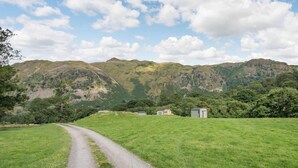 Ländliche Landschaft mit einem Schotterweg, der zu einem kleinen Gebäude führt, umgeben von grünen Feldern und Bergen.