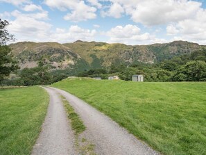 Paisagem rural com um caminho de cascalho que leva a um pequeno edifício, cercado por campos verdes e montanhas.
