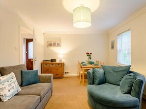 Ein Wohnzimmer mit einer Couch, einem Sessel, einem Esstisch mit Stühlen, einer Lampe und einem Fenster mit Jalousien.