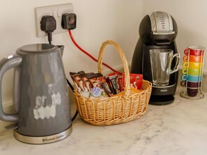 Eine Küchenarbeitsplatte mit einem Wasserkocher, einer Kaffeemaschine, einer Tasse und einer Korb mit Kaffeekapseln.