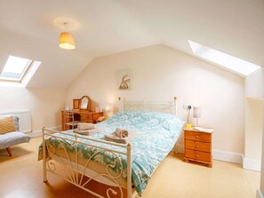 A bedroom with a sloped ceiling, a bed with a floral bedspread, a wooden dresser, a desk with a mirror, and a small bedside table with a lamp.
