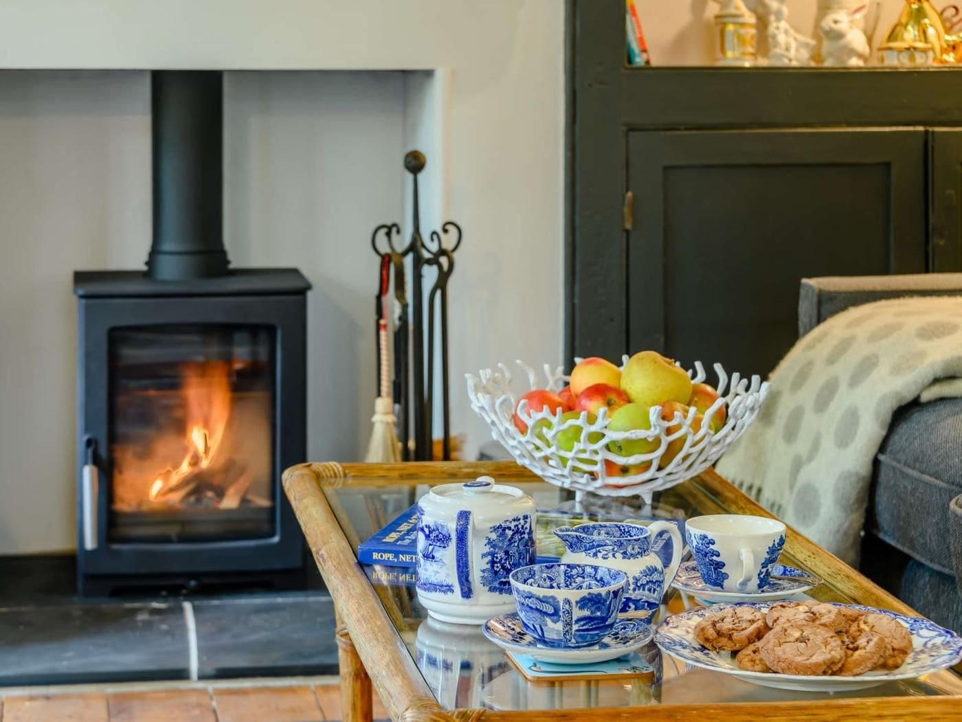 A cozy living room with a wood-burning stove, a fruit bowl, blue and white tea set, and a plate of cookies.