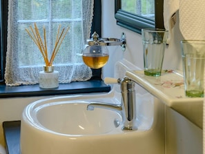 A bathroom sink with a chrome faucet, a glass of water, and a window with lace curtains.