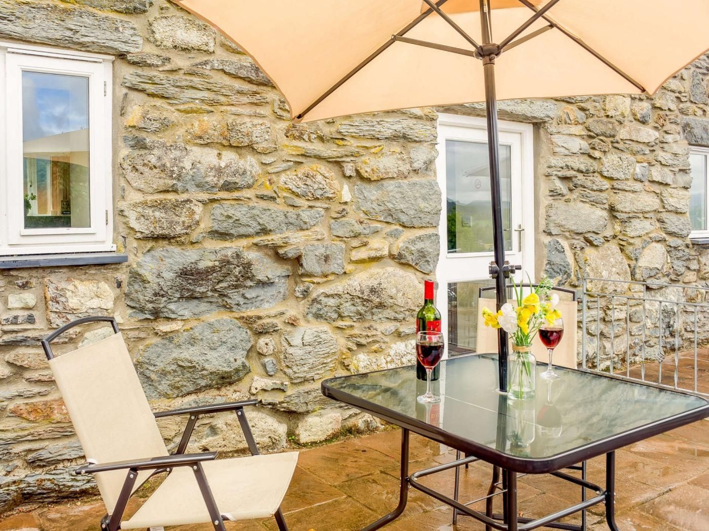 Esterno di un muro in pietra con un tavolo e una sedia in vetro, una bottiglia di vino e un vaso con fiori.