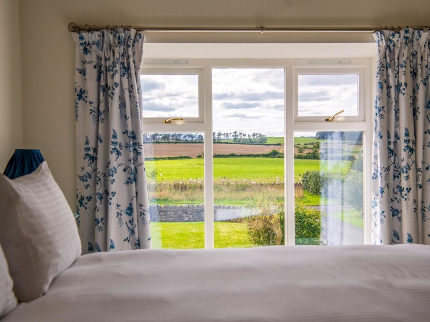 Une chambre à coucher avec un lit, des rideaux blancs ornés de motifs floraux bleus et une vue sur un champ verdoyant, visible à travers une fenêtre à cadre blanc.