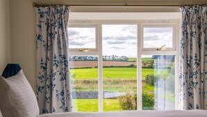 A bedroom with a bed, white curtains with blue floral patterns, and a view of a green field through a white-framed window. - 3 Bed in Belford (oc-89518) (Belford)