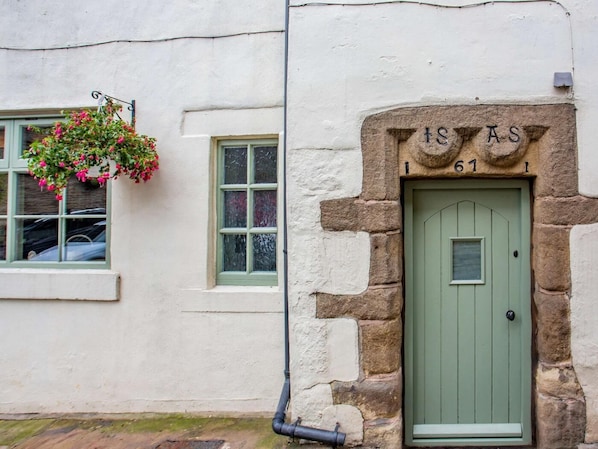 A traditional stone building with a green door, a window with a hanging flower basket, and a stone plaque above the door.