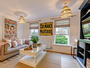A living room with a sofa, coffee table, and a television. There are two large windows with curtains, a hanging light fixture, and a sign on the wall. - 1 Bed in Cirencester (oc-90128) (Cirencester)