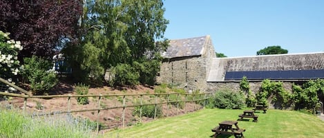 A stone building with a sloped roof, surrounded by a grassy area with lavender plants and picnic tables.