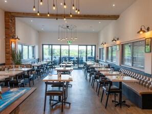 Tilava ravintola, jossa on useita pöytiä ja erillisiä istuinalueita, suuret ikkunat, joista on näkymä ulos, sekä erilaisia riippuvalaisimia ja seinään kiinnitettyjä valaisimia.