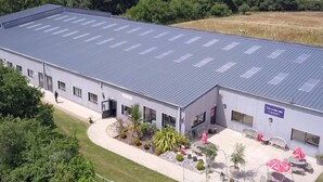 A large industrial building with a flat roof, a paved entrance, and a small outdoor seating area with umbrellas. - 3 Bed in Newport (oc-90245) (Newport)