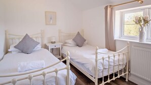 Un dormitorio con dos camas de metal blanco, una mesita de noche con lámpara, un radiador y una ventana con cortinas.