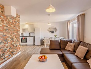 A modern living room with a brown leather sofa, a white coffee table with fruit, a kitchen in the background, and a dining area with a round table and chairs.