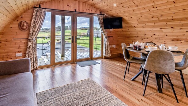 Uma cabana acolhedora com interior em madeira, uma área de refeições com cadeiras e mesa, e uma grande porta de correr de vidro que dá para um deck externo.