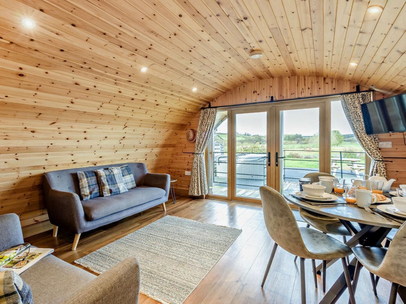 A cozy cabin interior with a wooden ceiling, a sofa, a dining table with chairs, and a large sliding glass door leading to an outdoor area.