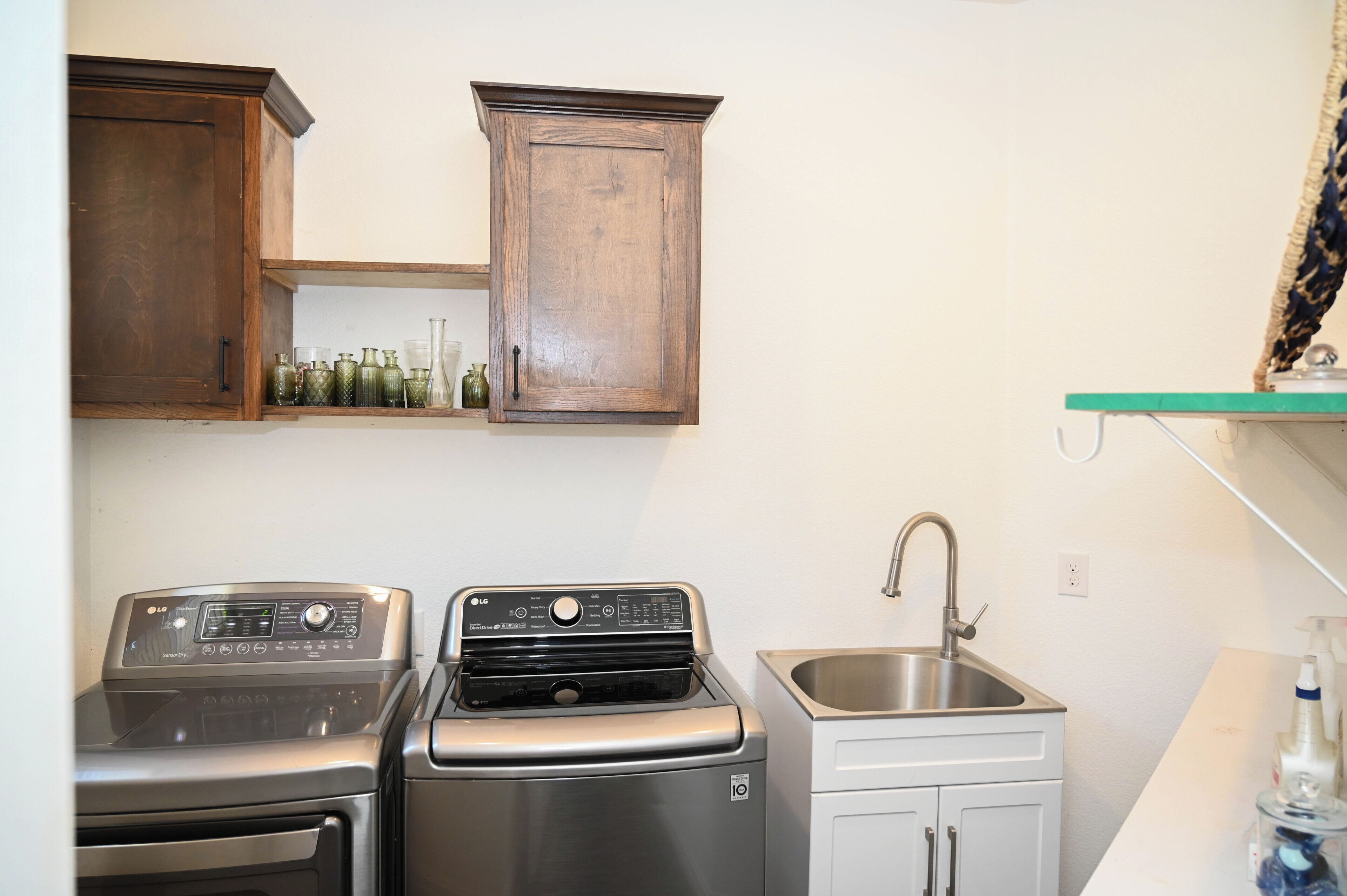 A laundry area with two washers and a sink, wooden cabinets above, and a shelf with bottles.