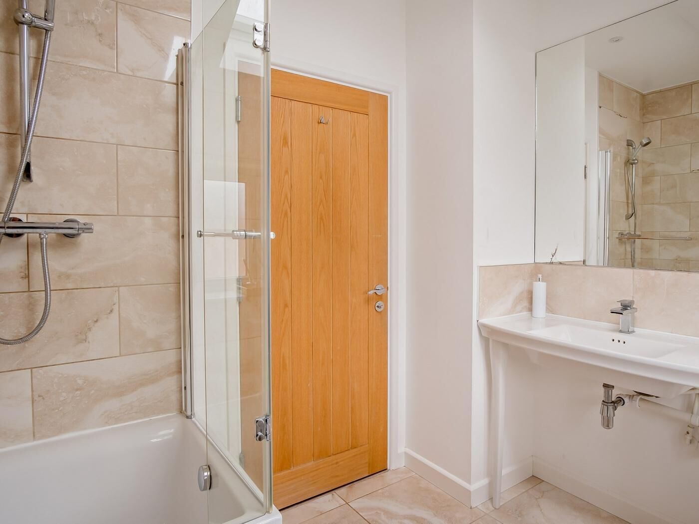 A bathroom with a glass-enclosed shower, a wooden door, a white sink, and a mirror.