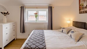 Um quarto com cama, mesa de cabeceira, abajur, janela com cortinas e um quadro na parede.