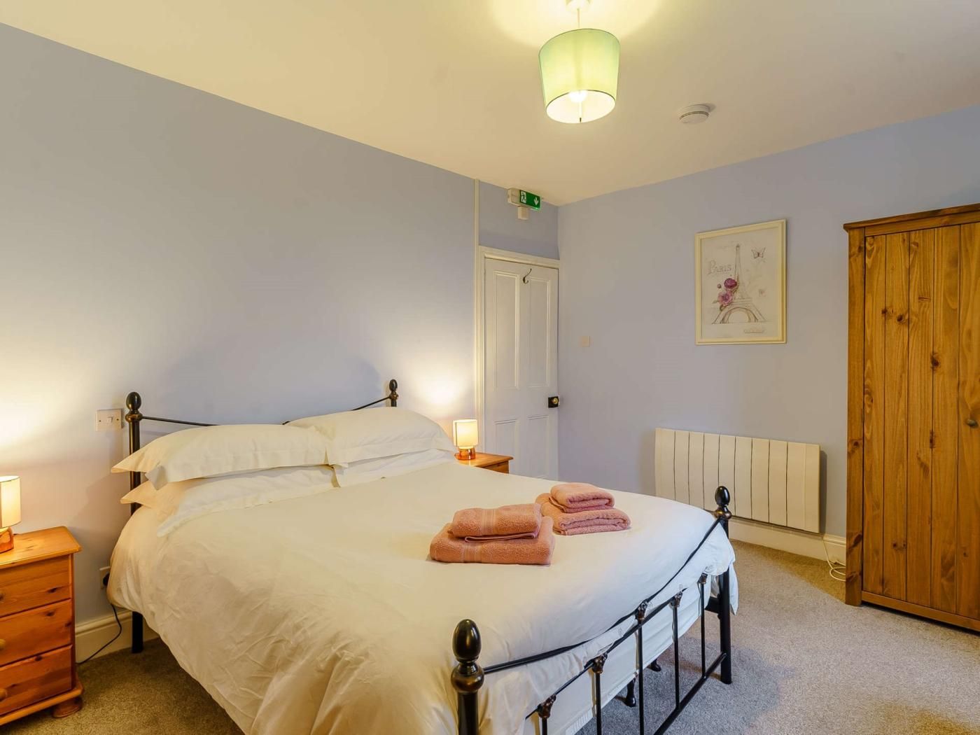 A bedroom with a bed, bedside tables, a radiator, a wooden wardrobe, and a door.