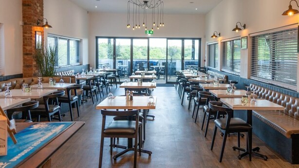 Un restaurant doté de nombreuses tables et de banquettes, d’un mur en briques et de grandes fenêtres.