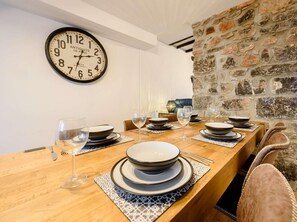 Ein Essbereich mit einem langen Holztisch, gedeckt für eine Mahlzeit, einer Wanduhr und einer Steinwand.