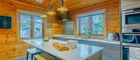 A modern kitchen with wooden paneling, a central island with stools, stainless steel appliances, and large windows.