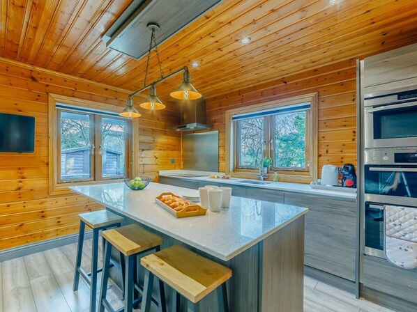 A modern kitchen with wooden paneling, a central island with stools, stainless steel appliances, and large windows.